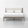 A modern wooden bed frame with a light upholstered headboard, made up with white bedding and two pillows, set against a plain white wall and floor.