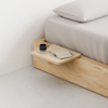 A minimalist wooden bedside table attached to a light wood bed frame holds a black coffee cup and saucer, along with a closed notebook and pen beside a neatly made bed with light bedding.