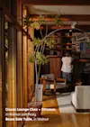 A man stands in a mid-century modern living room with wood and stone walls, arranging items on a shelf. Nearby are a potted plant, lounge chair with ottoman, side table, and sunlight streaming through large windows.
