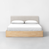 A modern bed with a light beige upholstered headboard, white bedding, and a wooden frame sits against a plain white wall on a light gray floor.