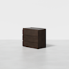 A dark brown, modern three-drawer dresser stands alone on a light gray floor against a plain white background.