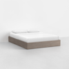 A minimalist bed with a light wood platform base, white mattress, white bedding, and two white pillows, set against a plain white wall in a simple, modern room.