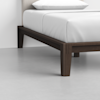 A close-up of a modern bed with dark wooden legs and a white mattress with white bedding, set on a light gray floor against a plain white wall.