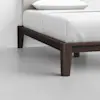 A close-up of a modern bed with dark wooden legs and a white mattress with white bedding, set on a light gray floor against a plain white wall.