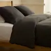 A minimal bedroom with a beige upholstered bed, white bedding, two pillows, a light green throw blanket, a brown nightstand with a modern lamp, and a beige rug on a tiled floor.