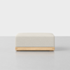 A square, light beige upholstered ottoman with a simple wooden base sits on a plain, light-colored floor against a white background.