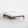 A minimalist wooden bed frame with a low profile, topped with a white mattress and two white pillows, set against a plain white background.