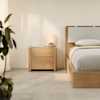 A modern, minimalist bedroom with a wooden bed and matching nightstand, topped with a small lamp, book, and cup. Soft natural light filters in, and a leafy plant is partially visible on the left.