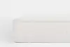 A close-up side view of a white mattress with a padded top and vertical stitched panels, set against a plain light background.