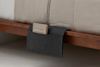 A felt bedside caddy in dark gray hangs over the side of a wooden bed frame, holding a small notebook with a beige cover and dark lines. The bed has beige and white bedding.