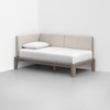 A minimalist daybed with a light wood frame and beige upholstered backrest, placed in the corner of a room with white walls and a gray floor. The bed has a white mattress and a single white pillow.