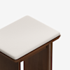 A close-up view of the corner of a wooden stool with a white cushioned seat, set against a plain white background.
