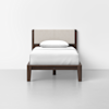 A single bed with a dark wooden frame, beige upholstered headboard, and white bedding with one white pillow, placed against a plain white wall in a minimalistic room.