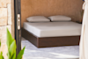 A neatly made bed with white sheets and two pillows sits on a dark wooden frame in a bright, minimalist room with beige walls and light carpeting. Green plants are visible in the foreground.