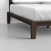 Close-up of a modern wooden bed frame with dark brown legs and white bedding, placed on a light-colored floor against a plain white wall.