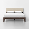 A modern bed with a dark wooden frame, beige upholstered headboard, and white bedding with two pillows, set against a plain white wall and light gray floor.