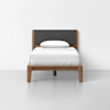 A minimalist single bed with a wooden frame, dark gray upholstered headboard, white mattress, and a white pillow, set against a plain white wall and floor.