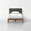 A minimalist single bed with a wooden frame, dark gray upholstered headboard, white mattress, and a white pillow, set against a plain white wall and floor.