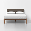 A modern bed with a wooden frame, dark upholstered headboard, white bedding, and two white pillows, set against a plain white wall and gray floor.