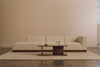 A minimalist living room featuring a beige sectional sofa, a wooden coffee table with a small vase and bowl, a light rug, and a large window allowing natural light into the space.