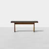 A minimalist wooden bench with a dark, cushioned seat and simple, straight legs, set against a plain white background.