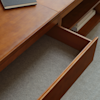 A wooden desk with an open drawer is shown on a gray carpet. On the desk surface, there is a closed black notebook. Books are stored on the lower shelf beneath the desk.