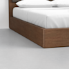 A close-up of a wooden bed frame with a white mattress, white sheets, and white pillows, set on a light gray floor against a plain white wall.