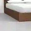 A close-up of a wooden bed frame with a white mattress, white sheets, and white pillows, set on a light gray floor against a plain white wall.