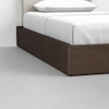 A close-up of a bed with a dark wooden platform frame, white bedding, and a white pillow, set on a light gray floor.