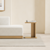 Minimalist living room with a light gray sofa, a wooden side table holding a cup and stacked books, and a neutral rug on a light floor, against a beige wall with part of a framed artwork visible.