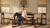 A woman in a tank top and wide-leg pants sits on a wooden bench at a dining table with matching chairs in a minimalist room with soft lighting and neutral decor.