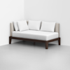 A modern daybed with a dark wooden frame, light upholstered backrest, and three white pillows, positioned in the corner of a minimalist room with white walls and a light gray floor.