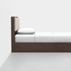 A minimalist wooden bed with a tall headboard, featuring a beige padded section and white bedding, positioned against a white wall and gray floor.
