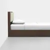 A minimalist wooden bed with a tall headboard, featuring a beige padded section and white bedding, positioned against a white wall and gray floor.
