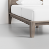 Close-up of a wooden bed frame with clean lines and a light gray finish, featuring white bedding and a pillow, set on a smooth light gray floor against a plain white wall.