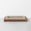 A minimalist beige pet bed with a cushioned fabric surface and a rectangular wooden frame, placed on a light gray floor against a plain white wall.
