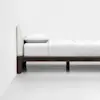 A minimalist wooden bed frame with a cushioned beige headboard, white bedding, and a single pillow against a plain white wall and floor.