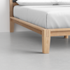 Close-up of the corner of a wooden bed frame with a light wood finish, showing part of a neatly made bed with white bedding and a smooth, light grey floor.