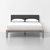 A modern bed with a wooden frame, dark gray upholstered headboard, white bedding, and two white pillows, set against a plain white wall and floor.
