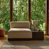A modern armless chair in ivory fabric sits beside a dark wood side table with a white lamp and a small plant. Large windows behind show lush green trees, filling the room with natural light.