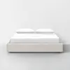 A modern bed with a beige upholstered frame and two white pillows, neatly made with white bedding, set against a plain white wall on a light gray floor.