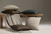 Four decorative pillows in neutral shades (beige, brown, and gray) are arranged on and around a wooden bench, with a textured beige blanket draped over the bench. The background is plain and softly lit.