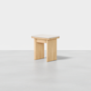 A small, minimalist wooden stool with three wide legs and a flat rectangular seat, set against a plain light gray background.