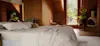 A cozy bedroom with a neatly made bed and white bedding, surrounded by warm wood walls. The room has green plants, a fireplace, and large windows letting in natural light.