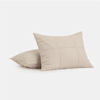 Two beige square pillows with subtle stitched grid patterns are stacked on a plain white background. One pillow leans against the other, both appearing soft and plush.