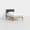 A minimalist twin bed with a light wooden frame, black upholstered headboard, and white bedding with a pillow, placed in a plain, empty room with light gray flooring and white walls.