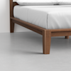 Close-up of the corner of a wooden bed frame with tapered legs, topped with a white mattress and pillows, on a smooth gray floor.