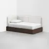 A modern daybed with a dark wooden frame and white cushioned backrest sits in the corner of a minimalist room with white walls and a light gray floor. A single white pillow rests on the mattress.