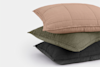 Three quilted pillows stacked on top of each other, with the top pillow in light pink, the middle in olive green, and the bottom in charcoal gray, against a plain white background.