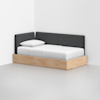 A minimalist daybed with a light wooden frame, white bedding, and a single white pillow. The backrest and side panels are upholstered in dark gray fabric, positioned against two white walls in a bright, empty room.